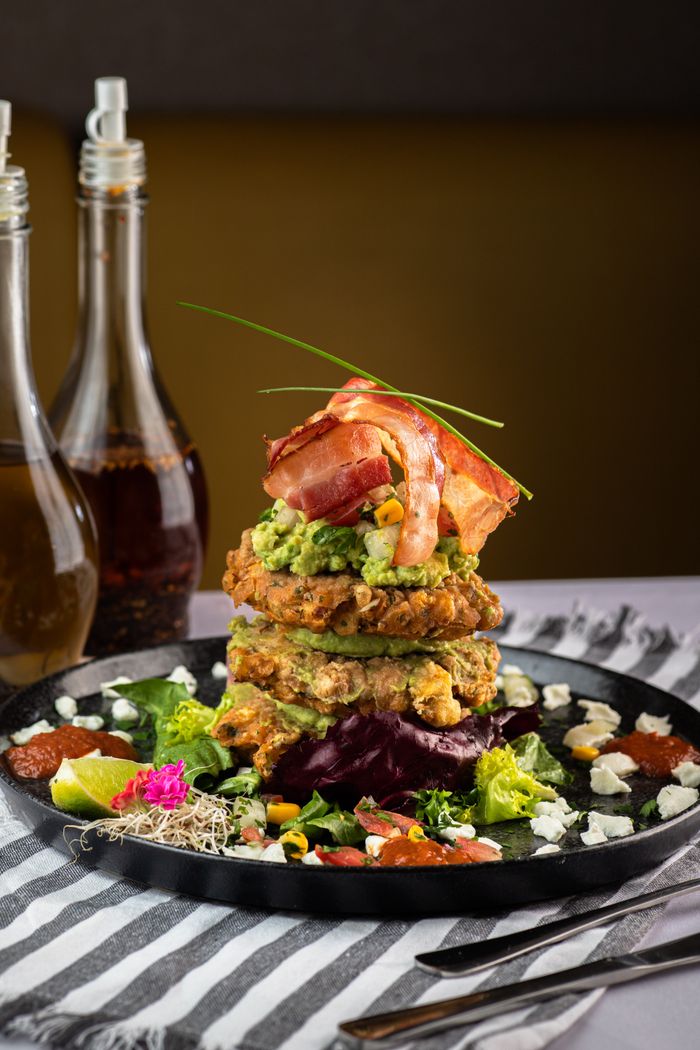 Enticing gourmet pancake stack with bacon, avocado, and vibrant salad on a black plate.